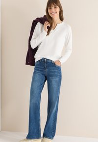 Haut blanc à manches longues avec un col henley, associé à un jean bleu foncé à jambes larges. Le mannequin tient une veste duveteuse violette. Fond clair.