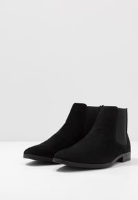 Botas Chelsea pretas em camurça com painéis laterais elásticos, bico fino, salto baixo e textura suave e matte. Design minimalista.