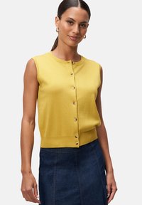 Cardigan sans manches jaune avec un col rond, fermeture à boutons à l'avant et une taille légèrement cintrée. Associé à une jupe en denim foncé.