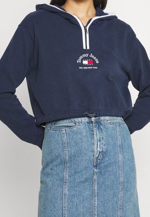 Granatowa krótka bluza z kapturem z półzamkiem i logo "Tommy Jeans", zestawiona z niebieską spódnicą jeansową z wysokim stanem.