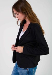 Svart kavaj med strukturerade slag, långa ärmar och smal passform, bärs över en vit skjorta, parat med blå jeans. Slät tygtextur.