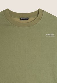 Camiseta de algodón verde oliva con cuello redondo. Presenta un pequeño logo blanco "FREDDY" y el texto "HOY ES TU DÍA" en la parte frontal. Textura suave.