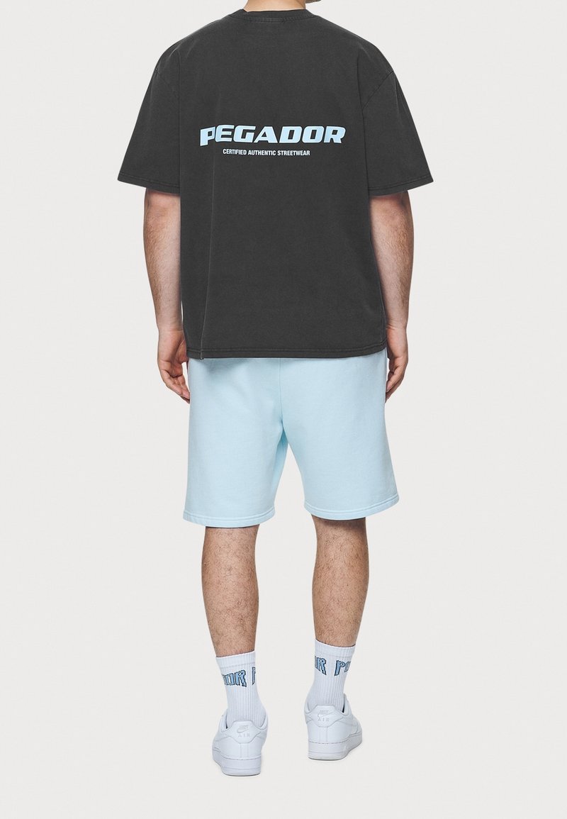Černé oversized tričko s modrým nápisem "PEGADOR", v kombinaci se světle modrými šortkami, bílými ponožkami a bílými teniskami, viděné ze zadní strany.