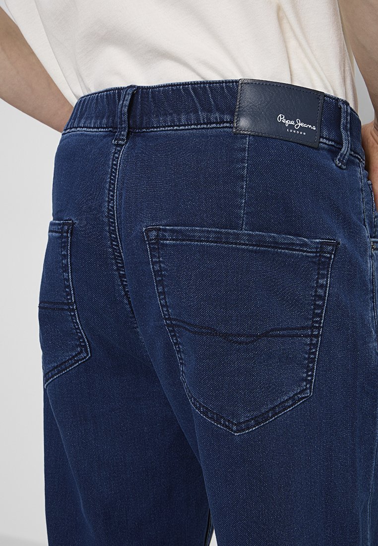 Jeans en denim bleu foncé avec une ceinture flexible, deux poches arrière et un patch en cuir avec la marque "Pepe Jeans London".
