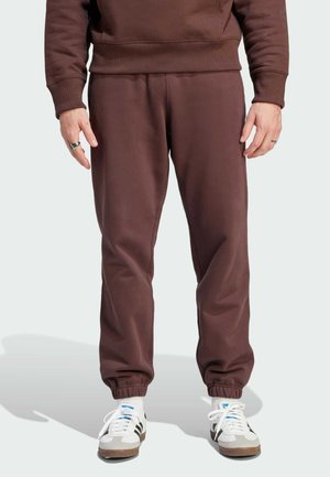 Pantaloni sportivi marroni con polsini elasticizzati, realizzati in tessuto misto cotone. Abbinati a sneakers bianche con accenti neri e blu.