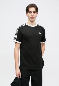 Čierne bavlnené tričko s bielym výstrihom a lemovaním rukávov, ktoré má na každom pleci tri biele pruhy a malé logo na hrudi.