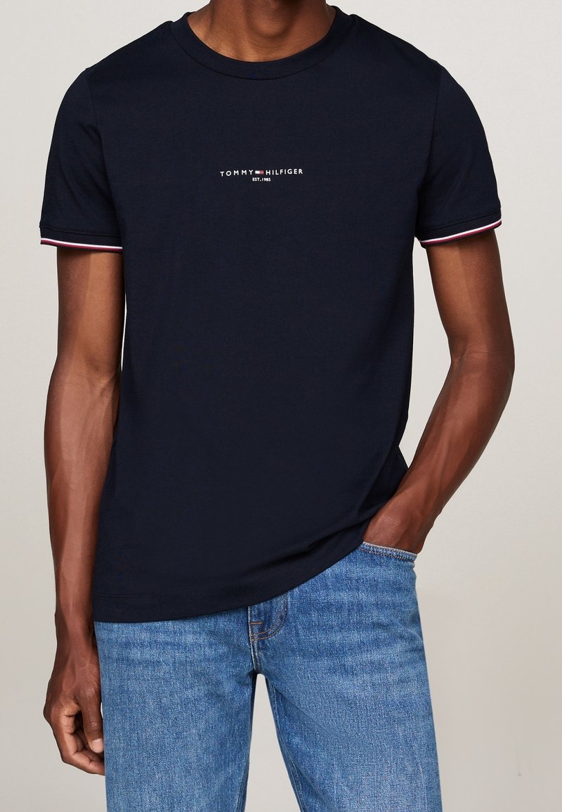 Navy blue short-sleeve t-shirt with a crew neck, featuring red and white striped accents on sleeves, and a white logo at the chest.