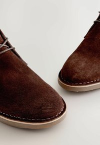 Gli stivaletti in suede marrone presentano una punta arrotondata, dettagli di cucitura sottili e una suola in gomma di colore chiaro. I lacci sono ben visibili sulla parte superiore.