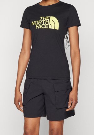 Person iført en sort T-shirt med korte ærmer med gul "The North Face"-logo og sorte cargoshorts med store forlommer.