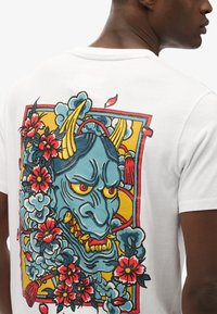 Camiseta blanca de algodón con un gráfico colorido de un rostro de demonio azul con ojos amarillos, rodeado de flores rojas y nubes estilizadas.