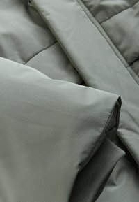 Jachetă padding gri, cu o carcasă exterioară netedă, design quilted și detalii din țesătură pliată, având un strat interior moale și texturat.