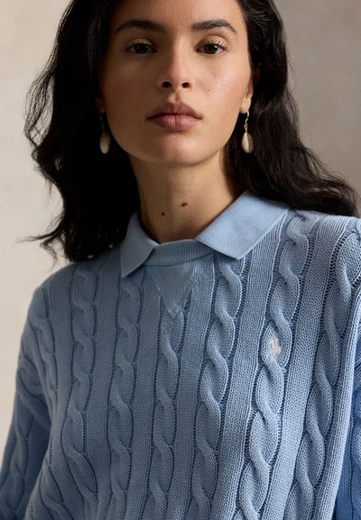 Młoda kobieta o ciemnych włosach nosi niebieski sweter w warkocze na koszuli z kołnierzykiem oraz perłowe kolczyki wiszące, stoi przodem z neutralnym wyrazem twarzy.