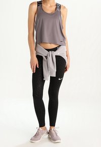 Camiseta sin mangas atlética gris con tirantes de logo negro de Nike; leggings negros con un patrón texturizado; sudadera gris claro atada a la cintura; zapatillas ligeras.