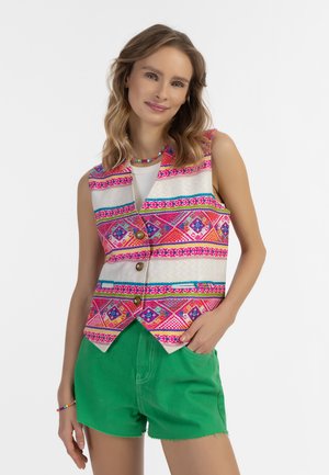 Vrouw met golvend haar draagt een kleurrijk patroon gilet over een witte top en groene korte broek, glimlachend met één hand in de zak tegen een witte achtergrond.