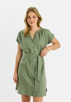 Joven mujer con cabello rubio hasta los hombros, vestida con un vestido verde oliva abotonado, de manga corta y con cinturón, de pie frente a un fondo liso.