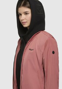 Veste bomber rose avec un sweat à capuche noir zippé en dessous. Comprend un logo brodé et un bouton-pression noir sur la manche, texture lisse.