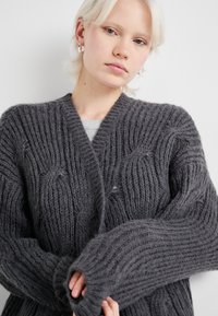 Grauer gestrickter Cardigan mit einem strukturierten Rippmuster, tiefem V-Ausschnitt und oversized Ärmeln. Hergestellt aus weichem, flauschigem Stoff.