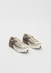 Zapatillas de baja altura hechas de materiales texturizados en gris y beige, con suelas de goma blanca, cordones y acentos de marca en el lateral.