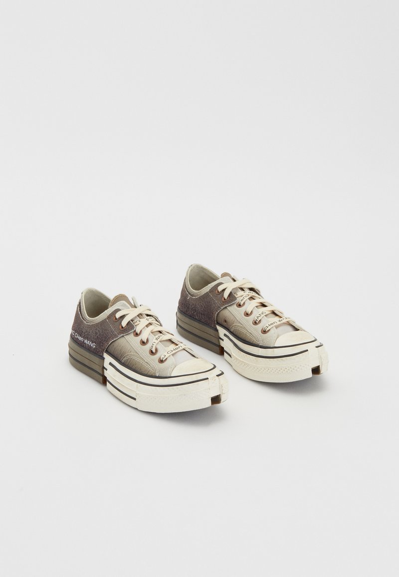 Zapatillas de baja altura hechas de materiales texturizados en gris y beige, con suelas de goma blanca, cordones y acentos de marca en el lateral.