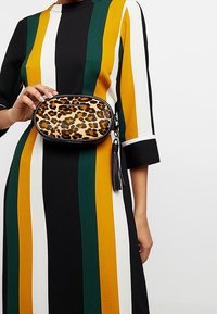 Oval väska med leopardmönster och svart kant, med dragkedja, accentuerad snodd och ett mönster av bruna fläckar på en ljus bakgrund.