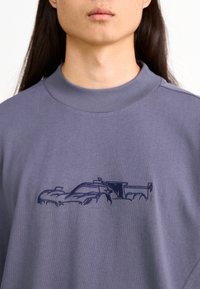 Sudadera gris con un cuello alto, que presenta un diseño de coche de carreras bordado en azul en el pecho. Textura de tela suave y lisa.