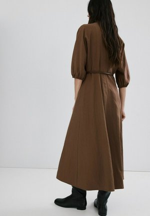 Žena s dugom tamnom kosom u smeđoj midi haljini s naduvanim rukavima i crnim gležnjačama, stoji uz jednostavnu svijetlu pozadinu.