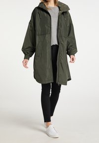 Chaqueta impermeable verde oliva con capucha, cierre de cremallera, puños elásticos y dos bolsillos delanteros, combinada con pantalones negros y zapatillas blancas.