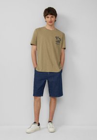 Khaki katoenen t-shirt met een blauwe grafische print, gecombineerd met donkerdenim shorts tot op de knie en witte sneakers met zwarte accenten.