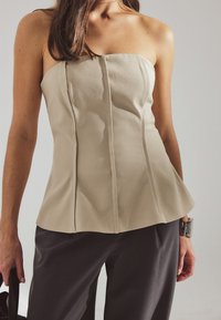 Woman wearing a beige strapless top and dark pants, holding a black handbag with a silver bracelet on her wrist against a neutral background.