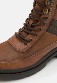 Bota de cuero marrón con paneles texturizados, ojales de metal, cordones marrones y suela de goma con un patrón de taco para estabilidad.