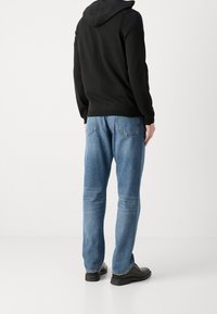 Schwarzer Kapuzenpullover mit lockerer Passform, kombiniert mit hellblauen Jeans im geraden Schnitt und mit Gesäßtaschen. Schwarze Schuhe.