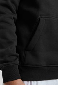 Felpa nera con tasca frontale, polsini e vita a coste, texture del tessuto liscia, mostra parte della manica e del corpo.