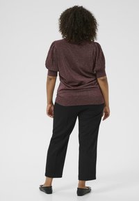 Woman with curly hair stands facing away, wearing a shimmery purple short-sleeve top, black cropped pants, and black flats.