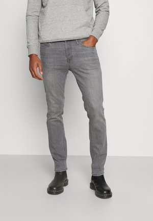 Homme portant un jean slim gris, un sweat-shirt gris clair et des bottines en cuir noir, debout avec une main dans la poche.