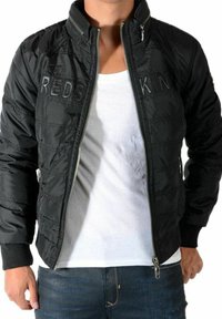 Sort dunjakke med ståkrave, lynlåslukning og frontlogo "REDSKIN." Har tekstureret stof og ribstrikkede manchetter.