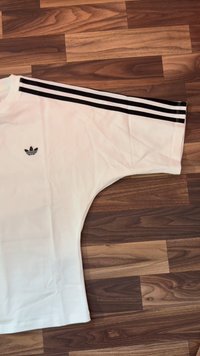 Λευκό κοντομάνικο μπλουζάκι Adidas με τρεις μαύρες ρίγες στον ώμο και μαύρο λογότυπο Adidas trefoil στο στήθος, τοποθετημένο επίπεδα σε ξύλινο πάτωμα.