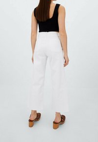 Jeans blancos de tiro alto y pierna ancha con dobladillo sin terminar, que cuentan con dos bolsillos traseros. Combinados con sandalias de plataforma marrones y una blusa sin mangas negra.