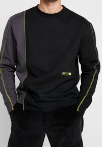 Sudadera negra y gris con paneles laterales en contraste, costuras amarillas y un pequeño logotipo. Cuenta con un cuello redondo y puños acanalados.