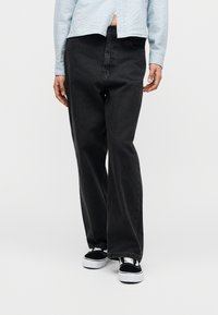 Μαύρο τζιν με φαρδύ μπατζάκι από τζιν ύφασμα, με κλασικό κούμπωμα κουμπιών και φερμουάρ. Συνδυάζεται με μαύρα και λευκά sneakers για αντίθεση.