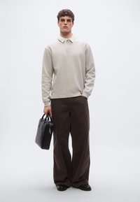 Tricou polo bej deschis cu mâneci lungi, pantaloni largi de catifea maro, geantă de mână din piele neagră, stând în fața unui fundal neutru.