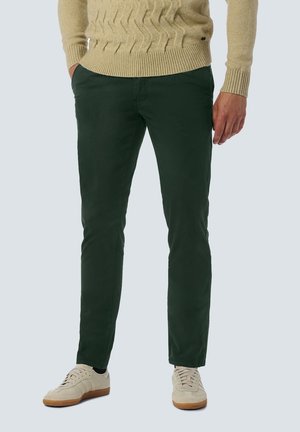 Mann trägt dunkelgrüne schmal geschnittene Hosen, beigen Strickpullover und weiße lässige Sneakers, steht mit einer Hand in der Tasche.