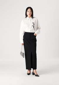 Cămașă albă cu guler, cu text negru, fustă neagră cu fante în față, tocuri negre strălucitoare și geantă mică din lanț argintiu. Design minimalist.