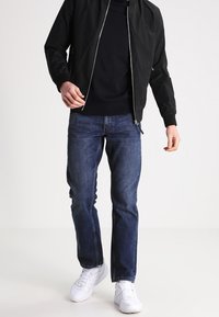 Černá bomber bunda se zipem, námořnický svetr, modré džíny a bílé tenisky. Bunda má klasický střih a hladký materiál.