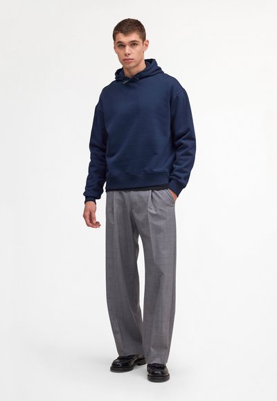 Junger Mann in einem dunkelblauen Kapuzenpullover, grauen plissierten Hosen mit weitem Bein und schwarzen Schuhen, der vor einem schlichten weißen Hintergrund steht.