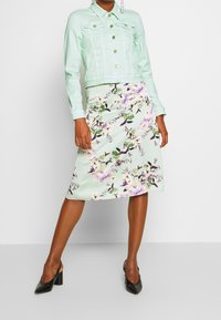 Miętowa dżinsowa kurtka ze złotymi guzikami, zestawiona z miętową spódnicą midi w kwiatowy wzór z fioletowymi i zielonymi akcentami. Czarne szpilki.