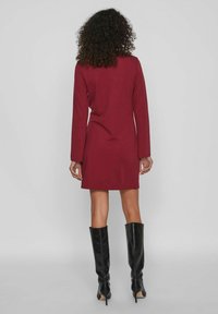 Vestido burdeos de manga larga con silueta ajustada, tejido suave y longitud a mitad del muslo, combinado con botas altas negras de tacón.