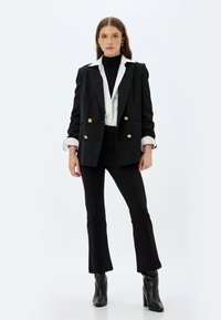 Musta mukautettu kaksirivinen bleiseri kultanapeilla, valkoisella kauluksella ja kapeilla mustilla housuilla, yhdistettyinä tiukkoihin mustiin nilkkasaappaisiin.