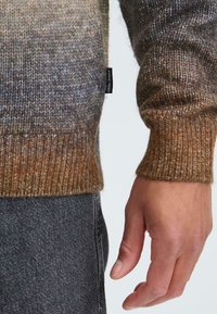 Suéter de punto en tonos marrones y grises degradados con puños de canalé. Presenta una pequeña etiqueta en el lateral. Combinado con jeans gris oscuro.