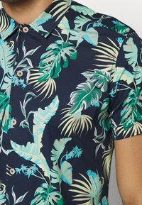 Košile s krátkým rukávem s tmavě námořnickým pozadím a velkými vzory tropických listů v zelené a světle modré. Dřevěné knoflíky na límci.