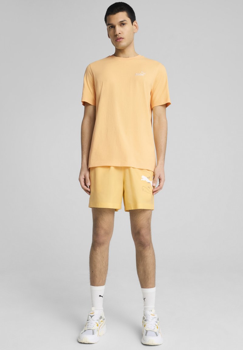 T-shirt di cotone arancione con un piccolo logo bianco, abbinato a pantaloni corti gialli con un logo bianco a motivo. Calze sportive bianche e scarpe completano il look.